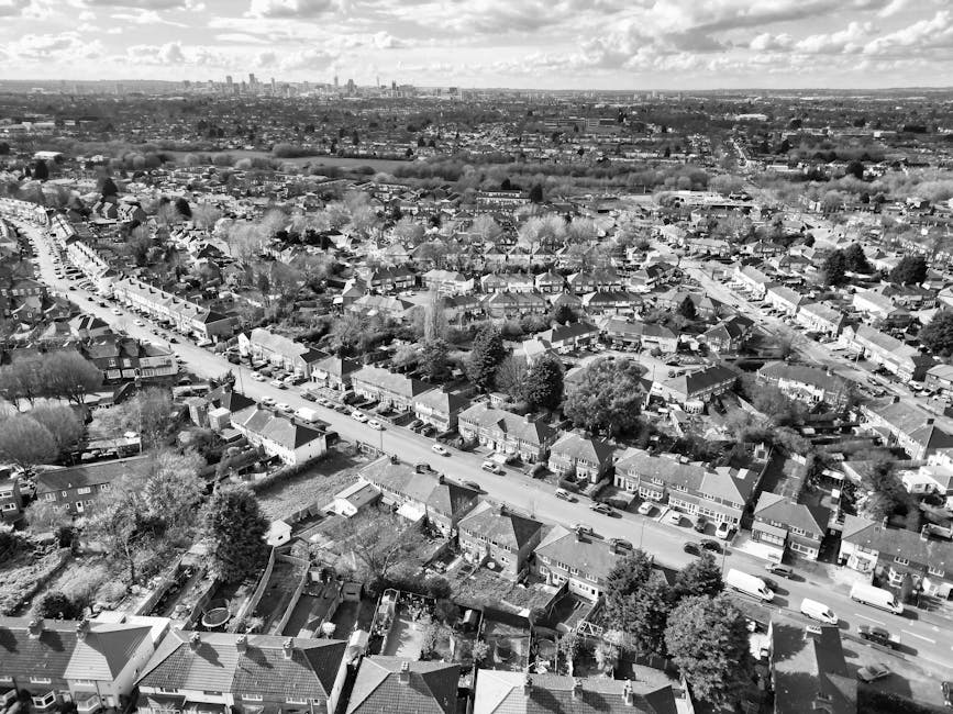 A black and white aerial photograph showcasing a residential neighbourhood with closely packed houses, each with distinct roofs made of tiles or shingles, and small gardens enclosed by fences. The ima