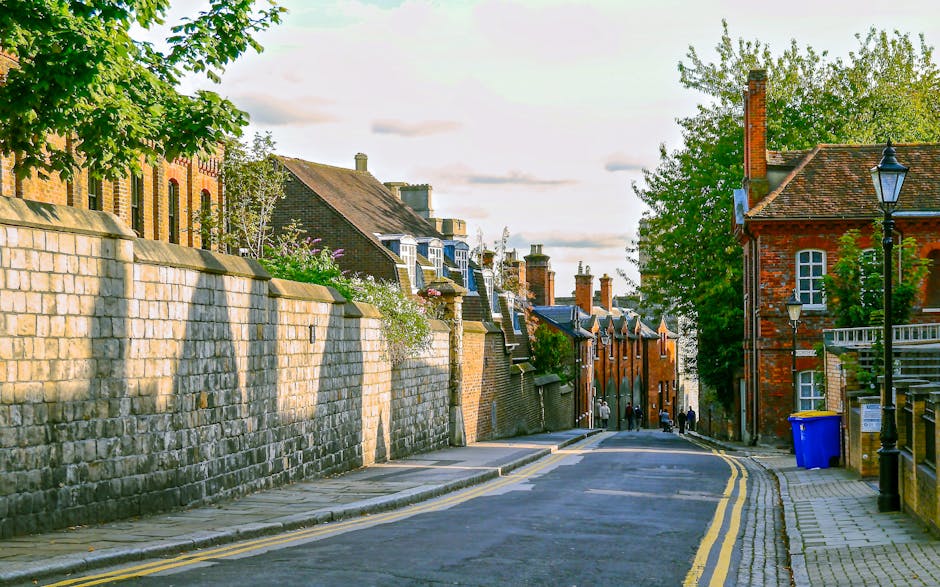 A quiet street scene in Enfield Town showing a narrow roadway with two yellow lines along the curb. On the left side, there is a high stone wall with trees and flowering shrubs behind it, while on the right side, brick buildings with pitched roofs and multiple chimneys line the street. A blue recycling bin is positioned near a lamp post beside the pavement, which has a few small groups of people walking in the distance. The pavement is wide and clean, with some area designated for loading or unloading. The sky is partly cloudy, with soft sunlight illuminating the scene, capturing the typical residential and historic character of Enfield. This setting is relevant for house removals and furniture transport, with potential for visualizing the process of home relocation or moving services provided by Removal Van Enfield, especially in areas with narrow streets and parking restrictions.