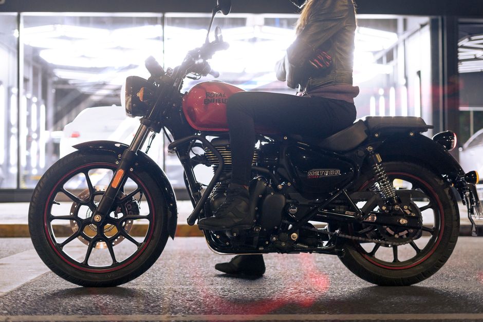 A person sitting on a black and red Royal Enfield Meteor motorcycle inside a parking garage or covered area with glass windows. The motorcycle is positioned parallel to the camera, showing the side profile, with visible components such as the front wheel with black rim and red accents, fuel tank, engine, and rear suspension. The person is wearing dark clothing and black boots, with their hands resting on the handlebars. The background features reflections of the interior lighting and parked cars through the windows. This scene illustrates the transportation aspect of house removals or vehicle relocation services provided by Removal Van Enfield, relevant to home relocation and transit of personal transport assets.