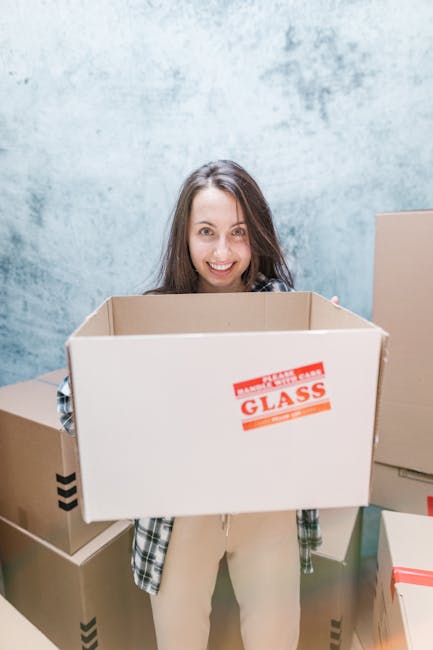 A woman with long brown hair and a smile is holding an open cardboard moving box in front of her, inside a room with a light blue textured wall. The box has a red and white label that reads 'Fragile Glass.' Several other packed cardboard boxes, some sealed with tape and others open, are visible around her, indicating a home packing or moving process. She is dressed in casual clothing, including a plaid shirt and light-colored trousers. The scene captures the packing stage of a house relocation, with the woman preparing items for safe transport. Removal Van Enfield’s professional removals service is suggested by the context, with the focus on packing, handling, and logistics involved in furniture transport and home moves.