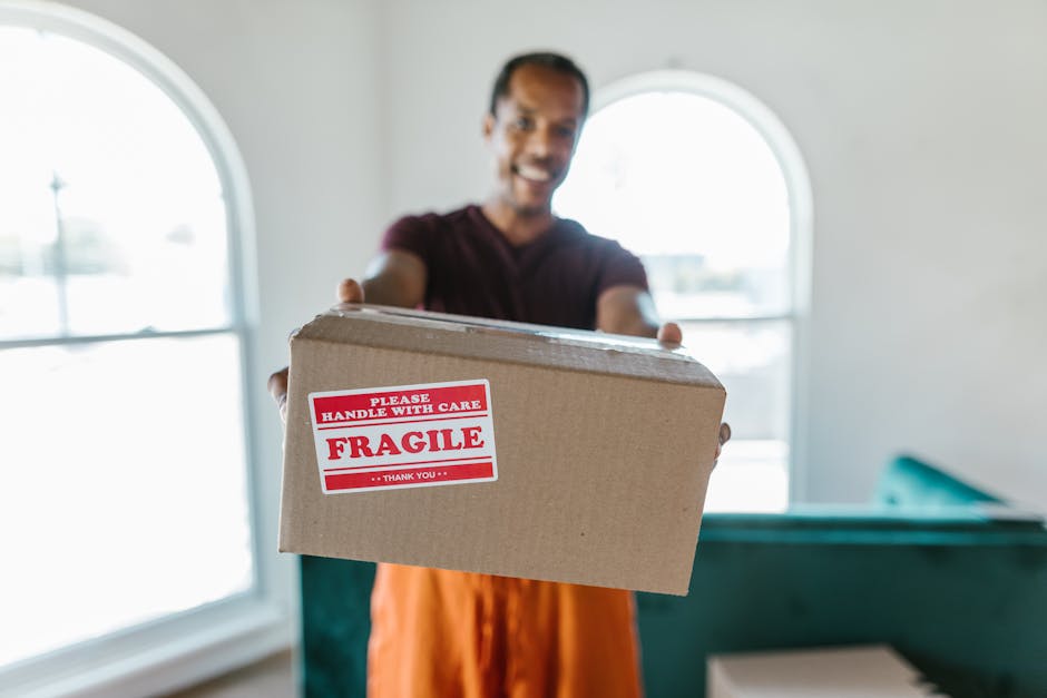 A man wearing a dark t-shirt and orange trousers is indoors, standing in front of large, arched windows with natural light streaming in. He is holding a medium-sized cardboard box towards the camera, which has a red and white warning label reading 'Please Handle with Care, Fragile, Thank You.' The interior setting includes a teal sofa and a white wall, indicating a home environment, possibly during a house relocation or packing process. The focus is on the box and the man’s outstretched hands, with the background slightly blurred. This scene illustrates the process of furniture transport and packing for a moving service, such as those offered by Removal Van Enfield, supporting themes of safe handling and home moving logistics.