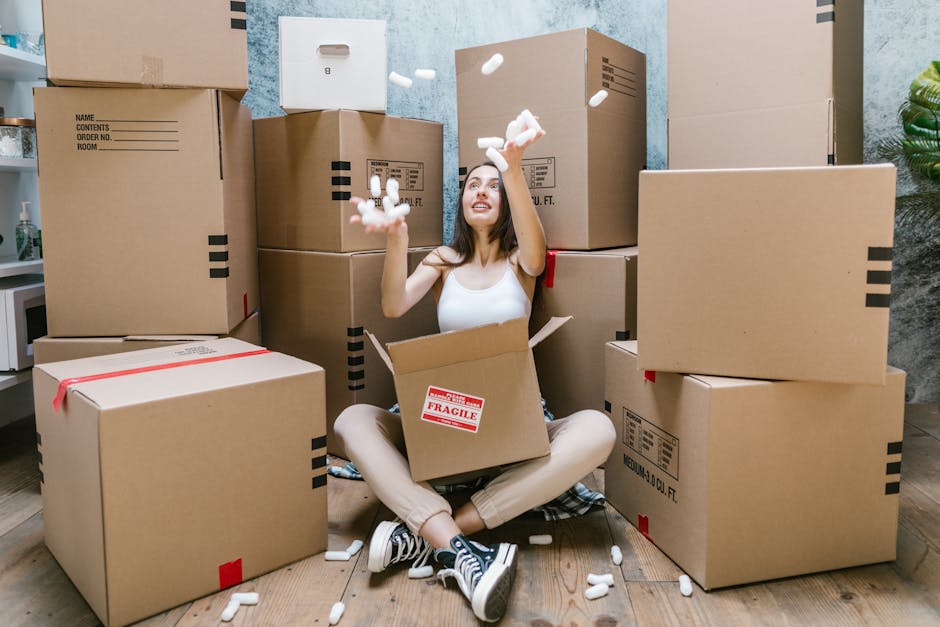 A woman with long brown hair and a smile is holding an open cardboard moving box in front of her, inside a room with a light blue textured wall. The box has a red and white label that reads 'Fragile Glass.' Several other packed cardboard boxes, some sealed with tape and others open, are visible around her, indicating a home packing or moving process. She is dressed in casual clothing, including a plaid shirt and light-colored trousers. The scene captures the packing stage of a house relocation, with the woman preparing items for safe transport. Removal Van Enfield’s professional removals service is suggested by the context, with the focus on packing, handling, and logistics involved in furniture transport and home moves.