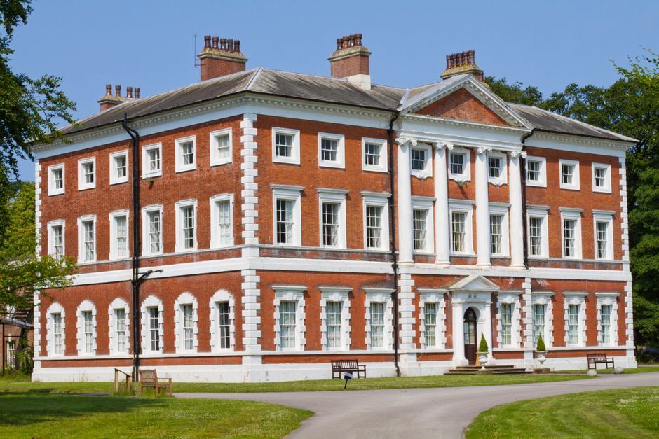 A large, historic red-brick building with white stone accents, situated on a well-maintained grassy lawn with a paved walkway in front. The building features multiple tall, white-framed sash windows arranged symmetrically on three floors, with decorative white cornices and a prominent central portico supported by four white columns. The roof is pitched with dark shingles and several brick chimneys protrude from the top. In the foreground, there are a few empty wooden benches along the walkway. The image appears to be taken during daytime with clear blue skies, and the surrounding greenery includes mature trees on either side of the building. This setting is suitable for visualising professional house removals involving large properties, with furniture and moving equipment possibly visible inside or near the entrance as part of a furniture transport or packing and moving process, highlighting the logistical aspects of home relocation services provided by Removal Van Enfield.