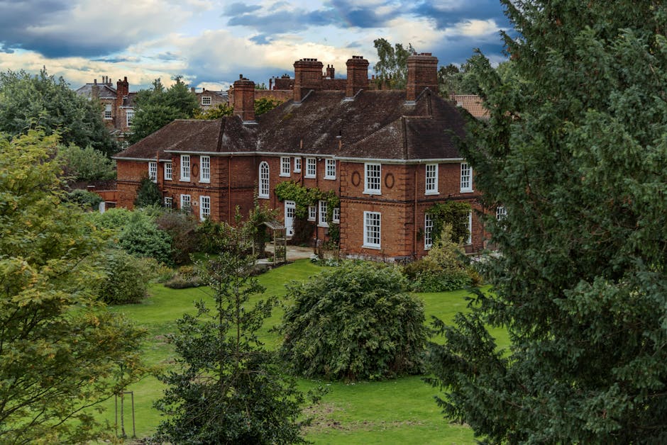 A large red-brick house with multiple chimneys, situated within a lush green garden. The property features white-framed sash windows and a wooden door partially visible behind dense foliage. The garden has well-maintained grass, mature trees, and various shrubs, with some trees partially obscuring the house. The scene appears to be set in the late afternoon or early evening, with diffused natural light and a partly cloudy sky overhead. In the foreground, there are large trees and bushes, framing the view of the house and its surrounding grounds. This setting suggests a quiet residential area, suitable for house removals or home relocation services, and is reminiscent of the type of large property that requires professional removals such as those offered by Removal Van Enfield, aligned with their expertise in house and furniture transport in the Enfield area.